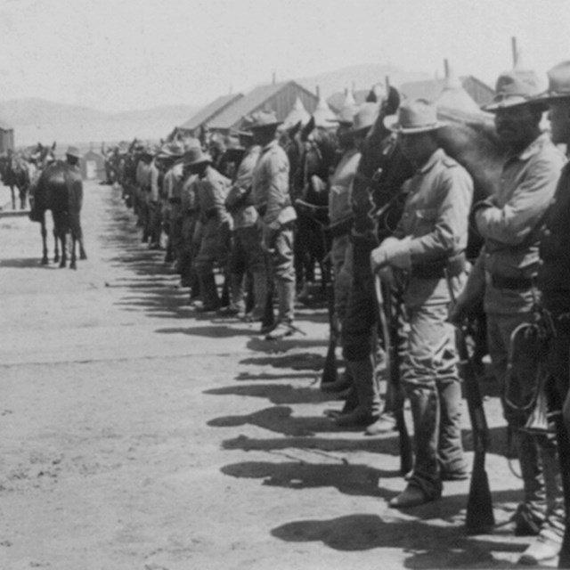 A line of Black soldiers standing in front of tents and wearing wide-brimmed hats