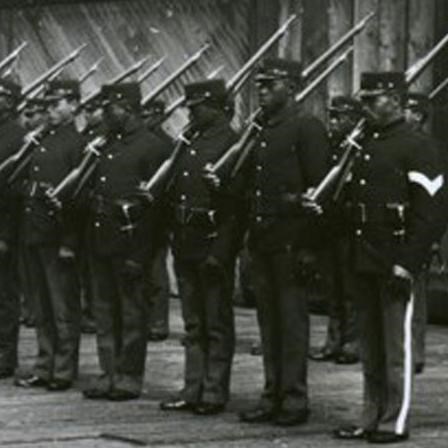 Several Black soldiers standing in a row on a wooden floor in front of two wooden buildings