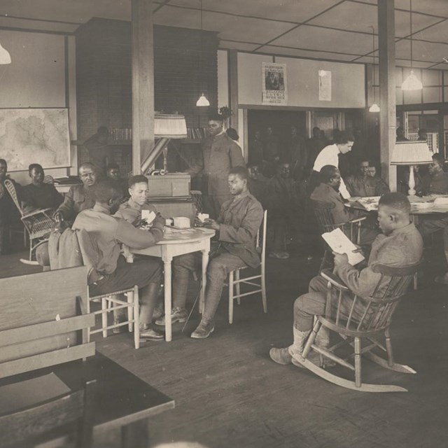 African American soldiers lounging in a room. Some reading and playing cards