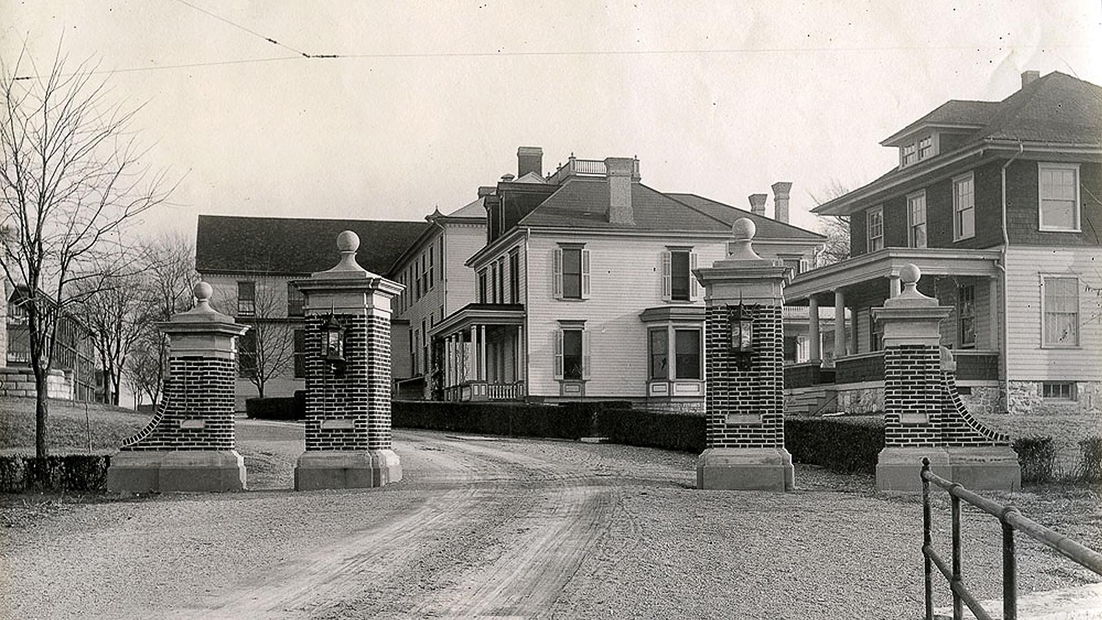 A dirt road with tire tracks leads towards two brick gateposts, and a row a two-story houses beyond.