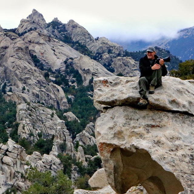 A man with a camera sits on a granite formation with spires rising from the surrounding mountains.