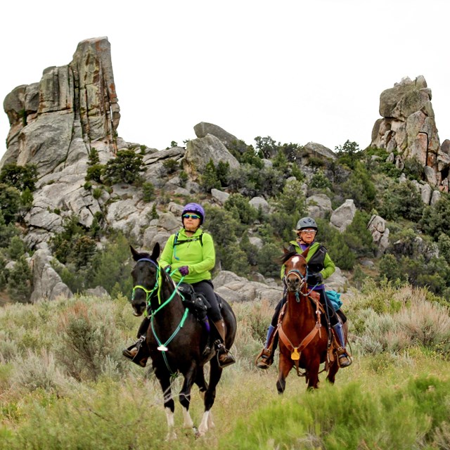 Two people on horseback ride down the trail in front of towering granite formations.