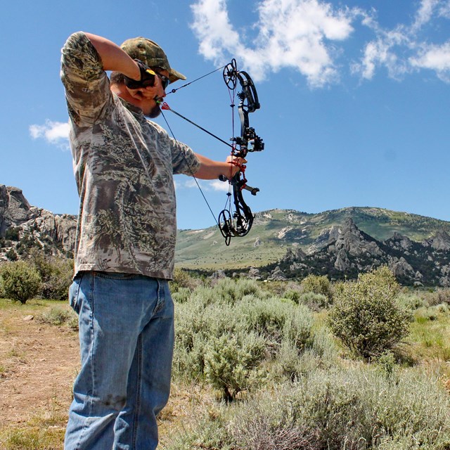 A man in a camo shirt draws back a compound bow aiming at a moose target in the distance.