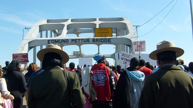 Rangers in flat hat mingle with large group walking across steel arch bridge