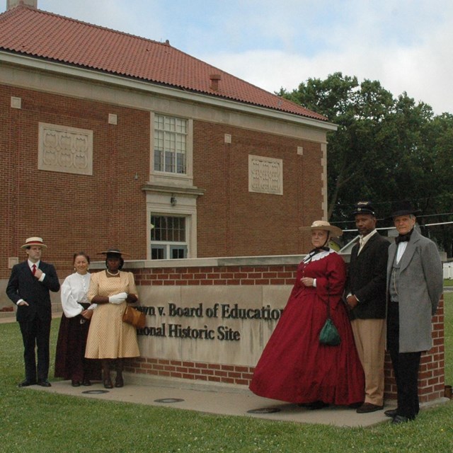 characters in period dress gather around park entrance sign, brick building in rear