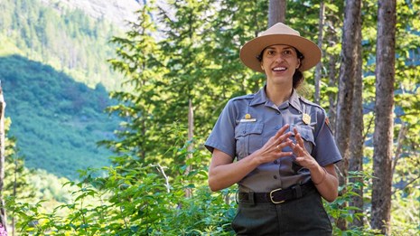 A park ranger speaks while smiling in front of green forest understory with view to mountainside