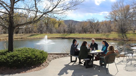 Several people sit at a picnic table in front of pond on sunny day