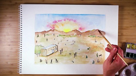 View of notebook paper with hand holding watercolor brush, painting a desert sunset image