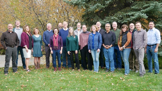 Photo of a couple dozen smiling people standing in grass in front of tree
