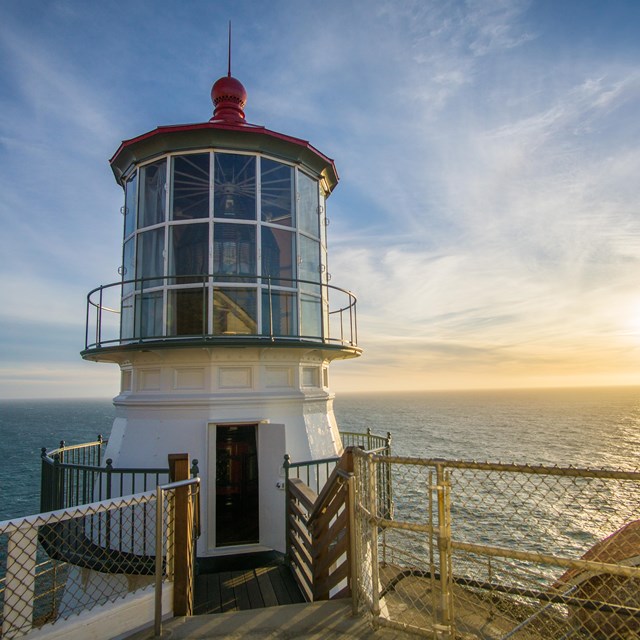 A closeup of a white lighthouse with blue ocean, wispy clouds, and setting sun behind