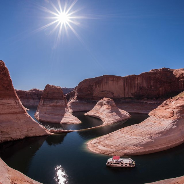 A glowing sun shines over red and white canyon with houseboat floating in low blue water