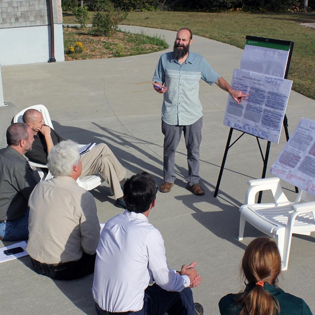 A man stands outdoors pointing to an easel with graphics while speaking to several seated people