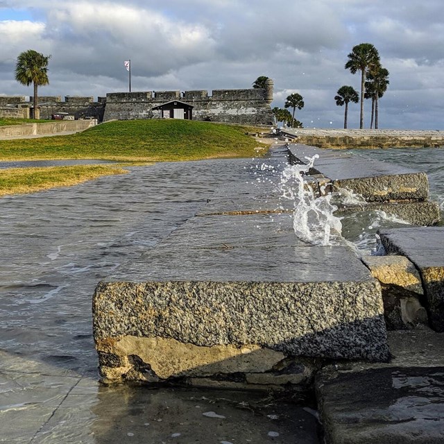 Ocean waves crash over a stone wall with palm trees and gray fort in background
