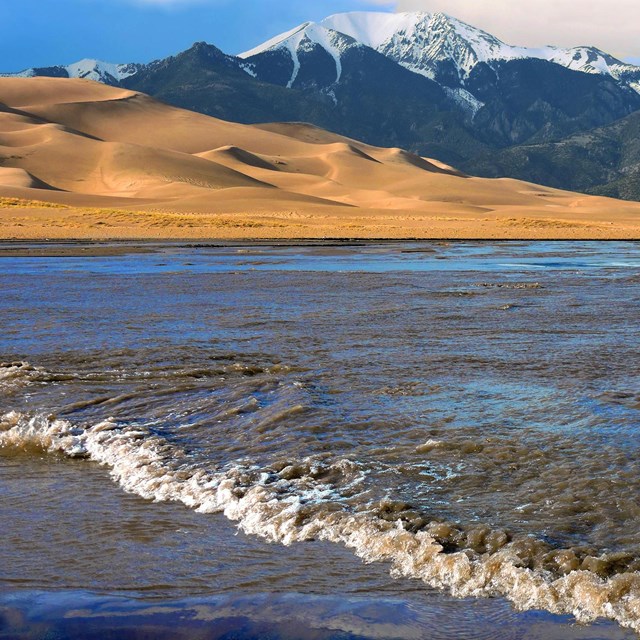 A stream in foreground with bright brown sane dunes and snowcapped mountains in distance