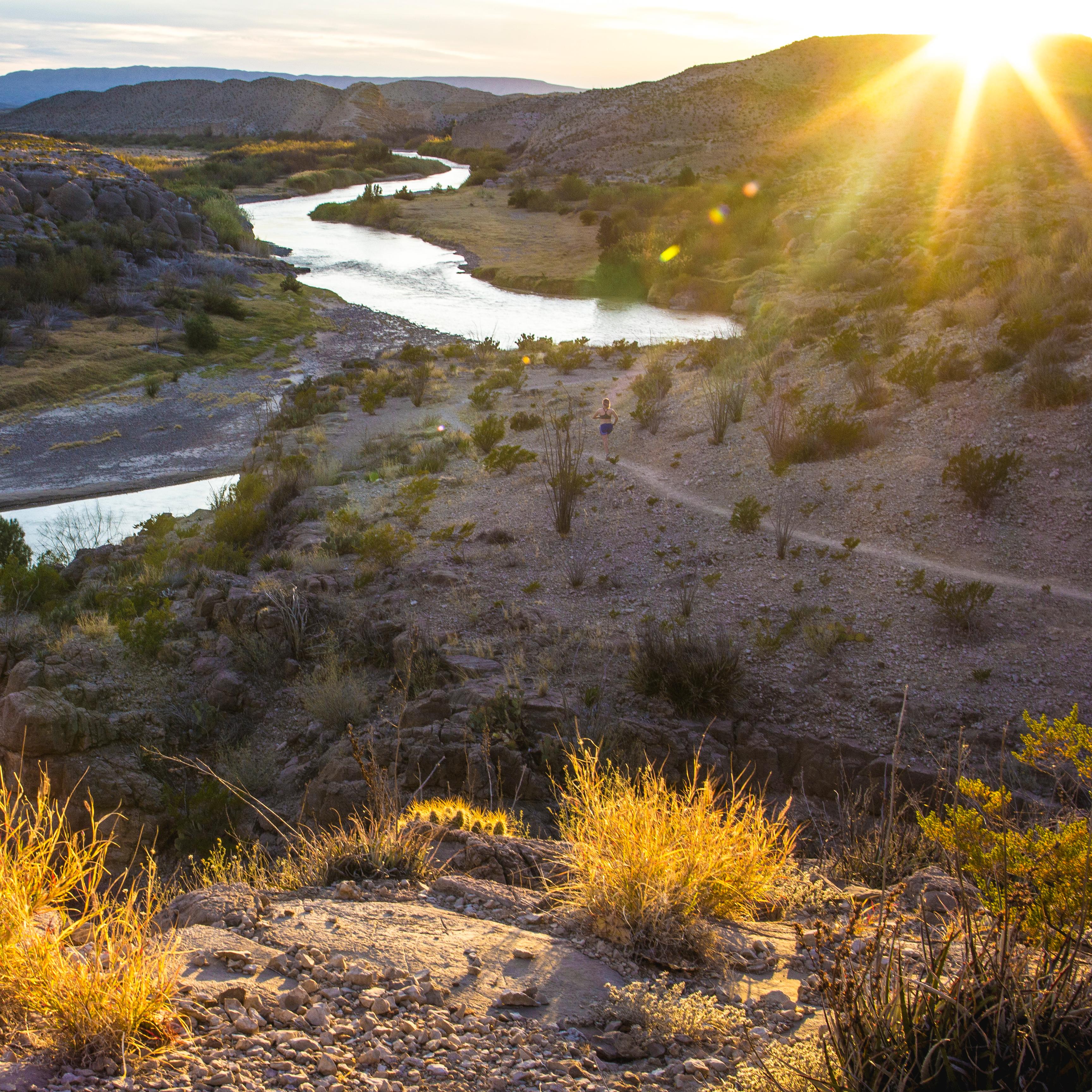 Sun sets behind desert hills with scrub and brush above a meandering river valley