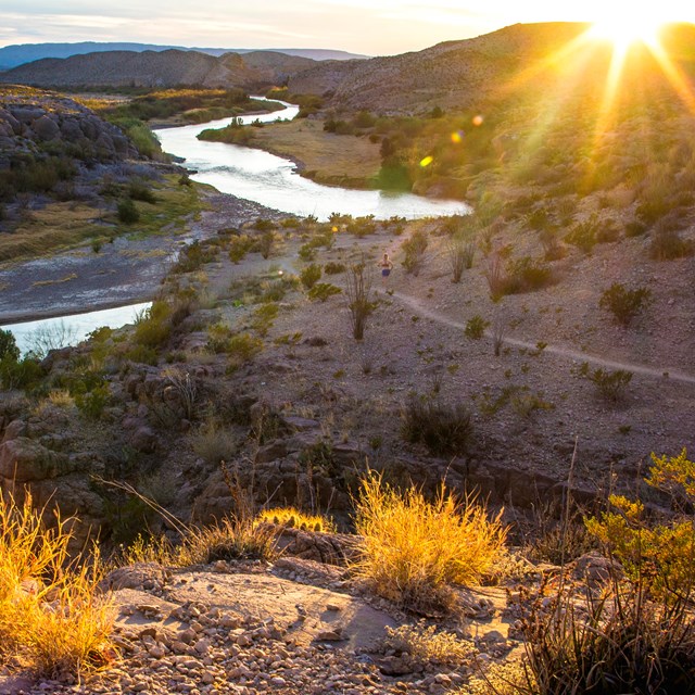 Sun sets behind desert hills with scrub and brush above a meandering river valley