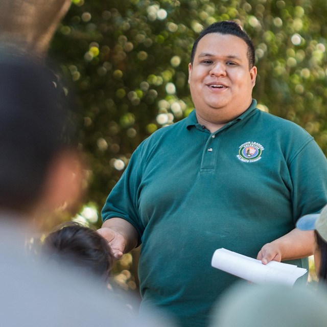 A young smiling intern speaks in front of trees while students in foreground listen