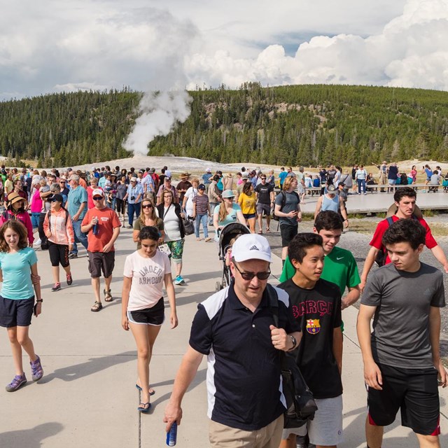 Geyser steams in distance as hundreds of people walk away on boardwalk toward camera