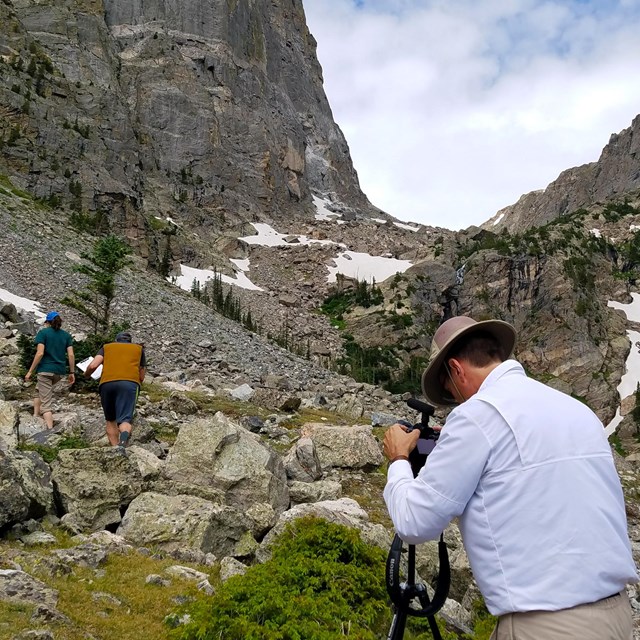 Man with video camera records a male and female walking through a boulder-filled mountain slope