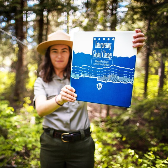 A park ranger in a forest holds up a binder titled 