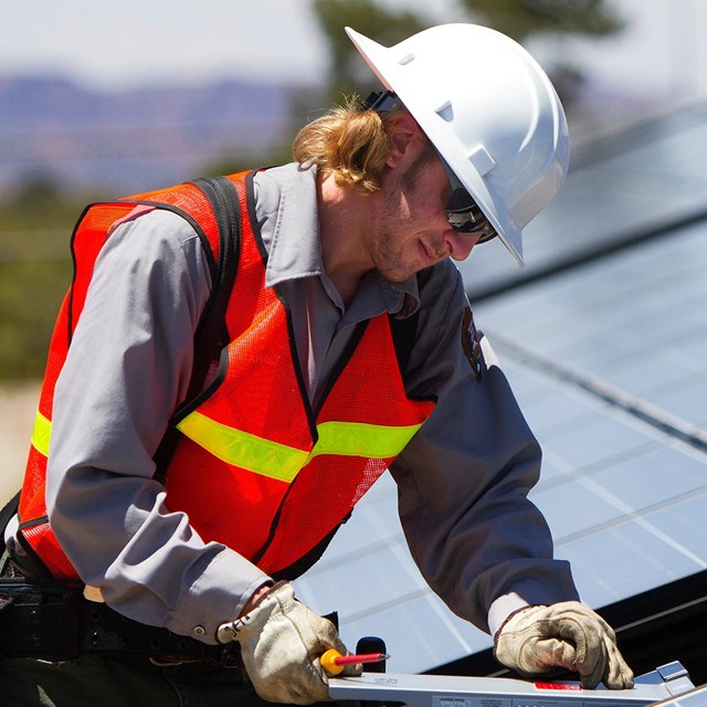 A maintenance employee in bright orange vest reviews a field of solar panels