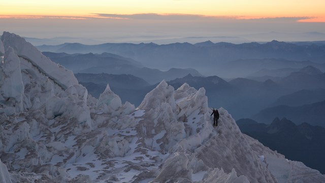 a person climbs along jagged peaks of ice with mountain ranges in the background during a yellow red