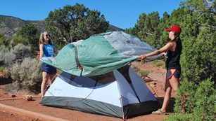 Two young women stretch a rain fly over a tent.