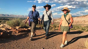 3 women wearing hiking clothing walking on a dirt trail silhouetted by blue sky.