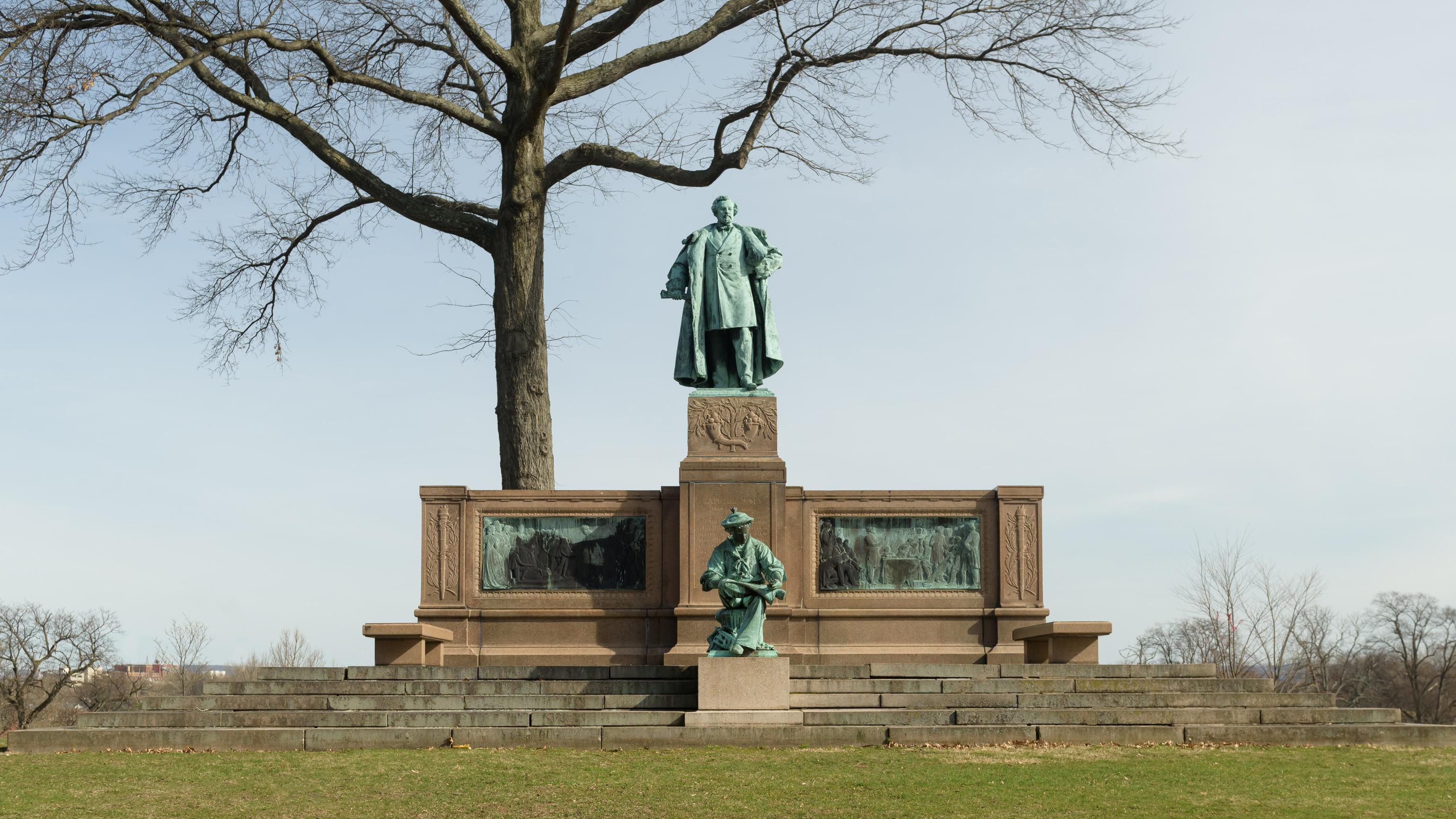A statue with two figures atop a granite steps, underneath a tree. 