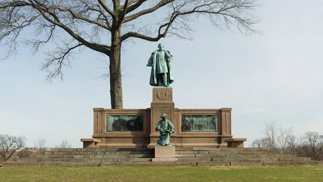 A statue with two figures atop a granite steps, underneath a tree. 