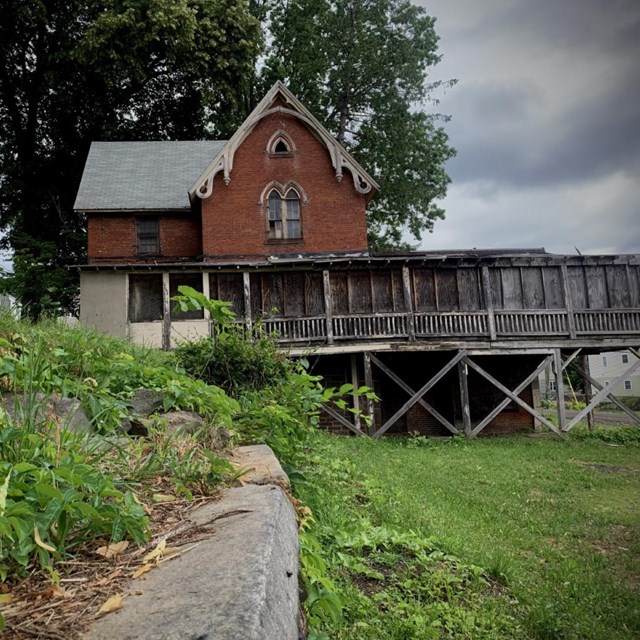 A two story red brick building with an enclosed porch underneath a tree. 