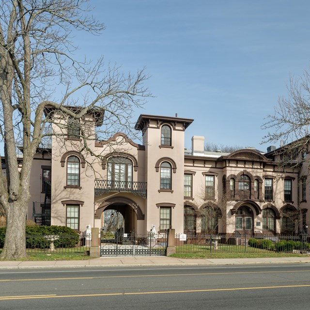 A multistory cream building with numerous windows trimmed in brown set behind trees. 