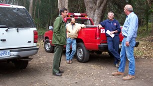 four men stand next to red pickup truck and white suv