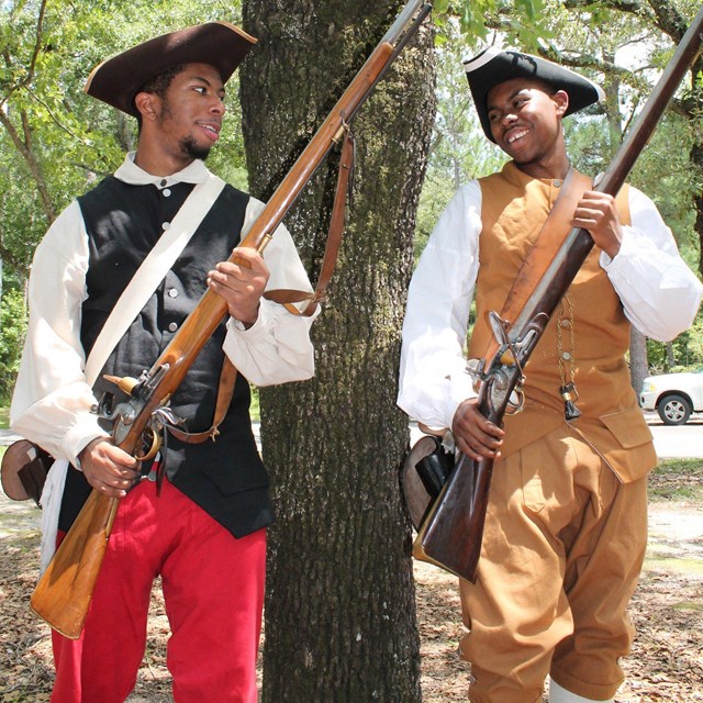 two men dressed as revolutionary soldiers hold muzzle loaders next to a tree