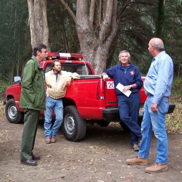 four men stand next to red pickup truck and white suv
