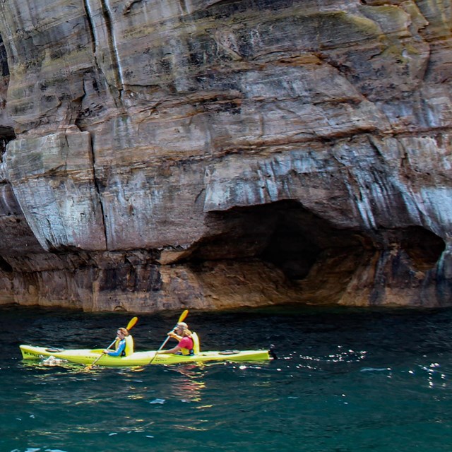 two people kayak in lake below huge rock face