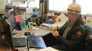 ranger in a green sweater and beige flat hat sitting at a desk holding a coyote skull