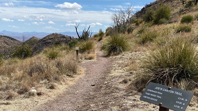 Trailhead sign with dirt trail, mountains, blue sky, and clouds in the distance.