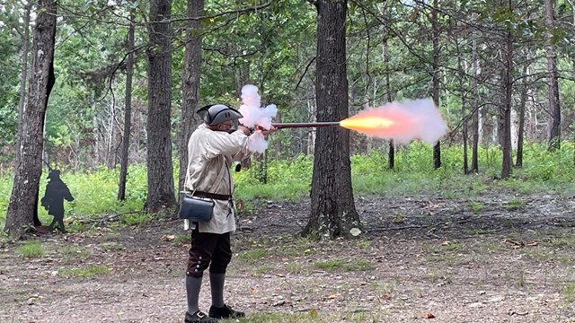 Reenactor in a tricorn hat and hunting frock fires a musket