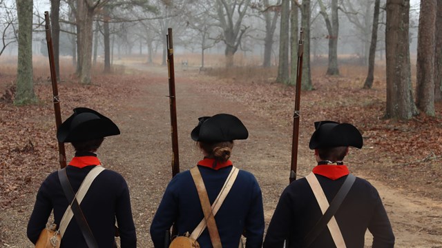 Three reenactors in Continental uniforms stand with muskets on a dirt road 