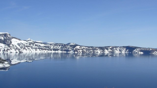snow covered caldera walls run horizontally between a cloudless blue sky and a deep blue lake