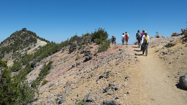 Hikers on a high elevation trail with sparce vegetation to Mount Scott Fire Lookout in the distance