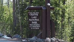Round wood logs painted dark brown support a brown wood sign stating the west entrance to the park