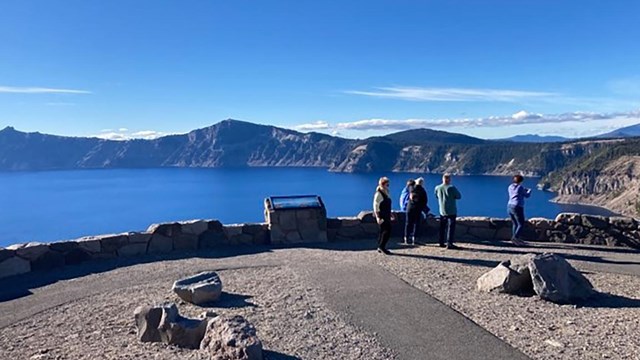 An exhibit set in a short stone wall at the end of a long sidewalk where people look at a blue lake.