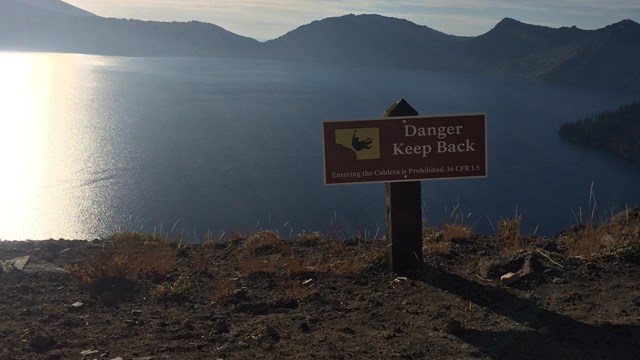 A wood sign with Danger painted in white is at the caldera edge with lake behind. 