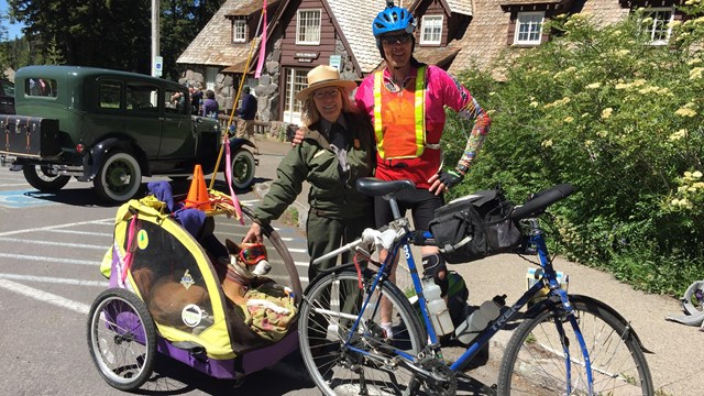 man and ranger behind a blue bicycle with a cart attached and a dog wearing goggles inside