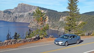 A car drives along on the East Rim Drive, with Crater Lake in the background.
