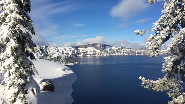 On left and right Snow-laden evergreens frame Crater Lake, the caldera rim and a partly cloudy sky
