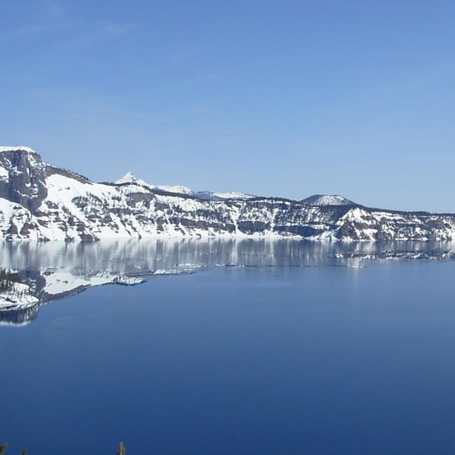 snow covered caldera walls run horizontally between a cloudless blue sky and a deep blue lake