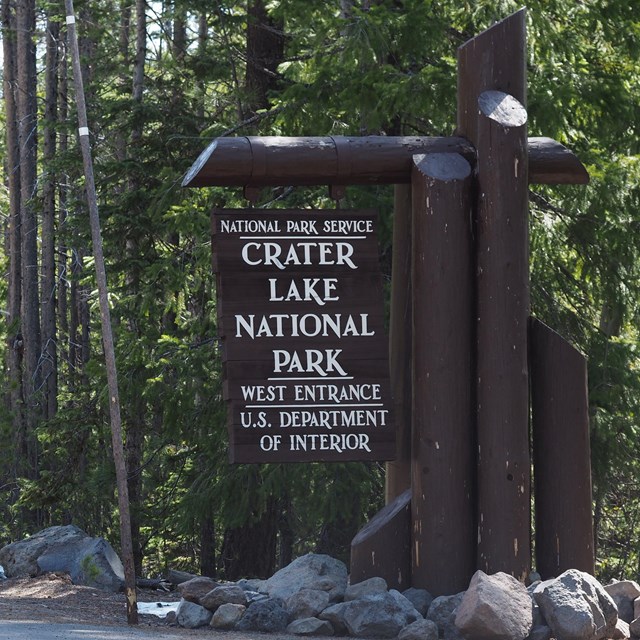 Round wood logs painted dark brown support a brown wood sign stating the west entrance to the park